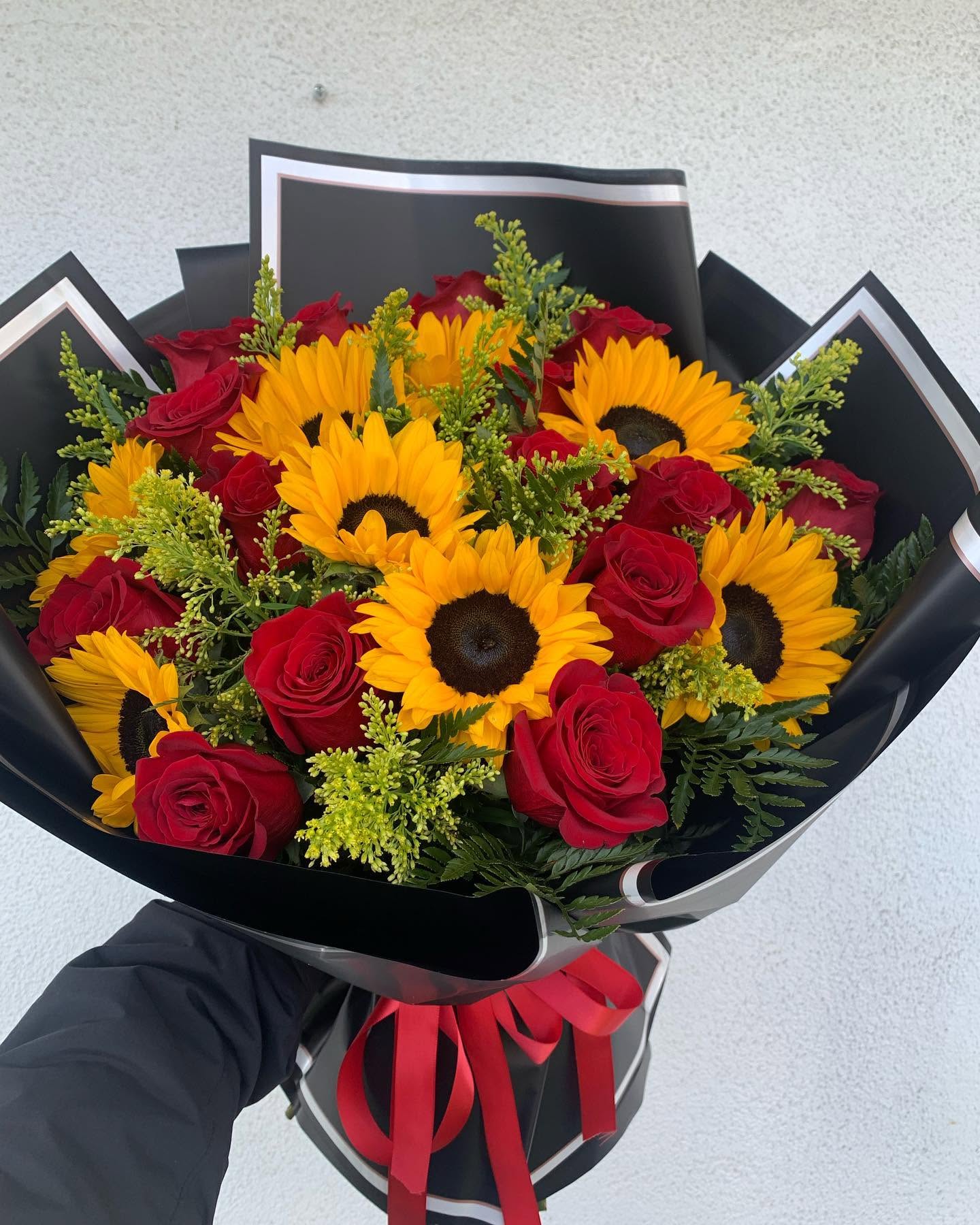 Red roses and bright yellow sunflowers bouquet with greenery, black wrapping, and red ribbon from The Hidden Gift Store
