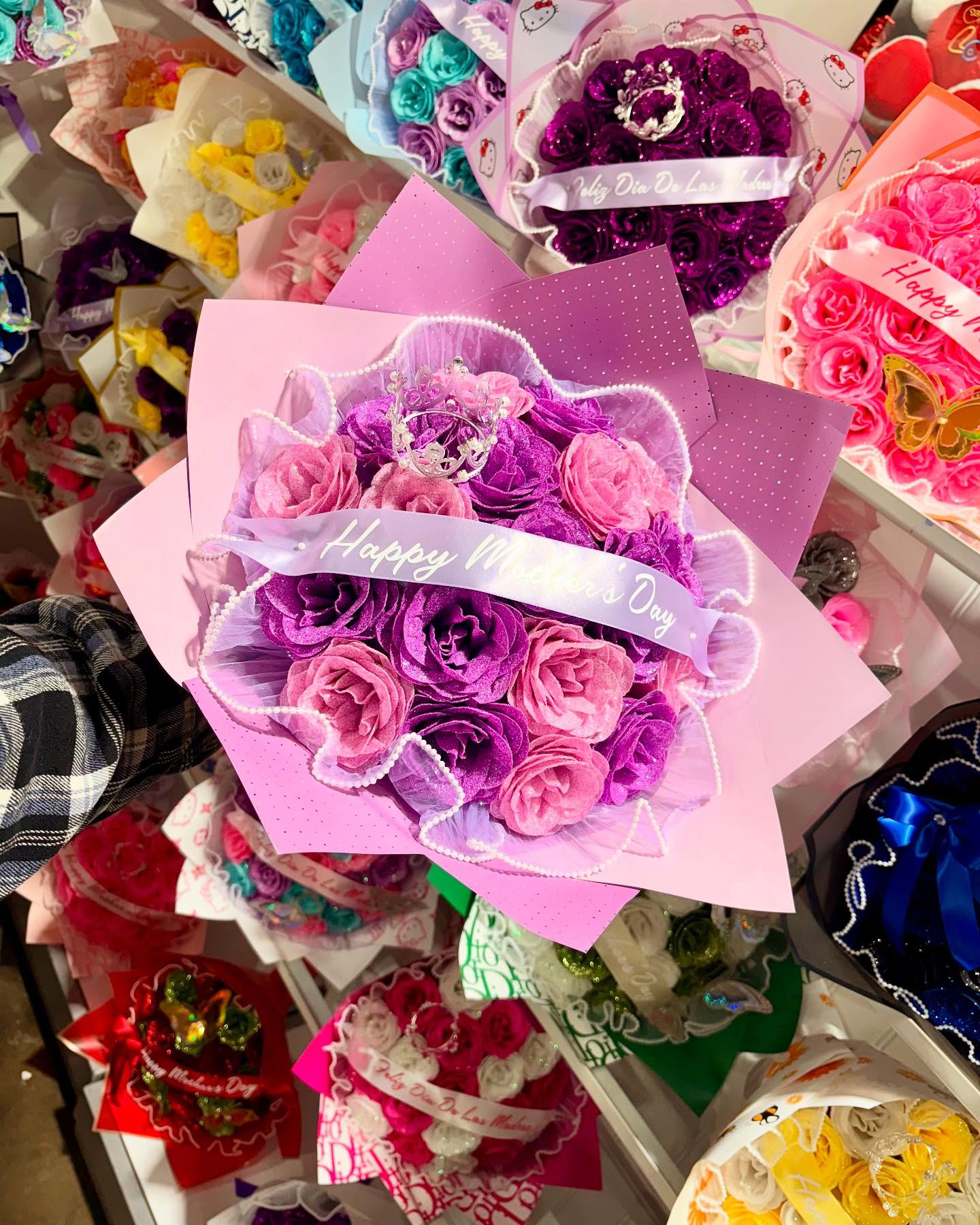 Pink and purple rose bouquet with a crown and “Happy Mother’s Day” ribbon, surrounded by colorful floral arrangements