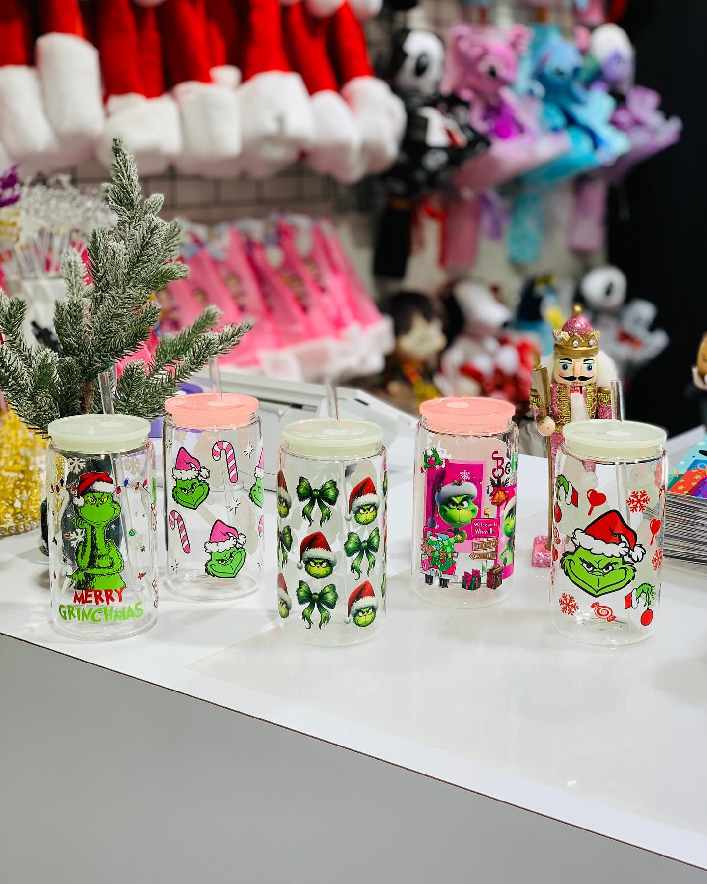 Five clear glass Christmas tumblers with lids and straws featuring festive Grinch designs and Merry Grinchmas text, displayed on a store counter with holiday decor in the background.