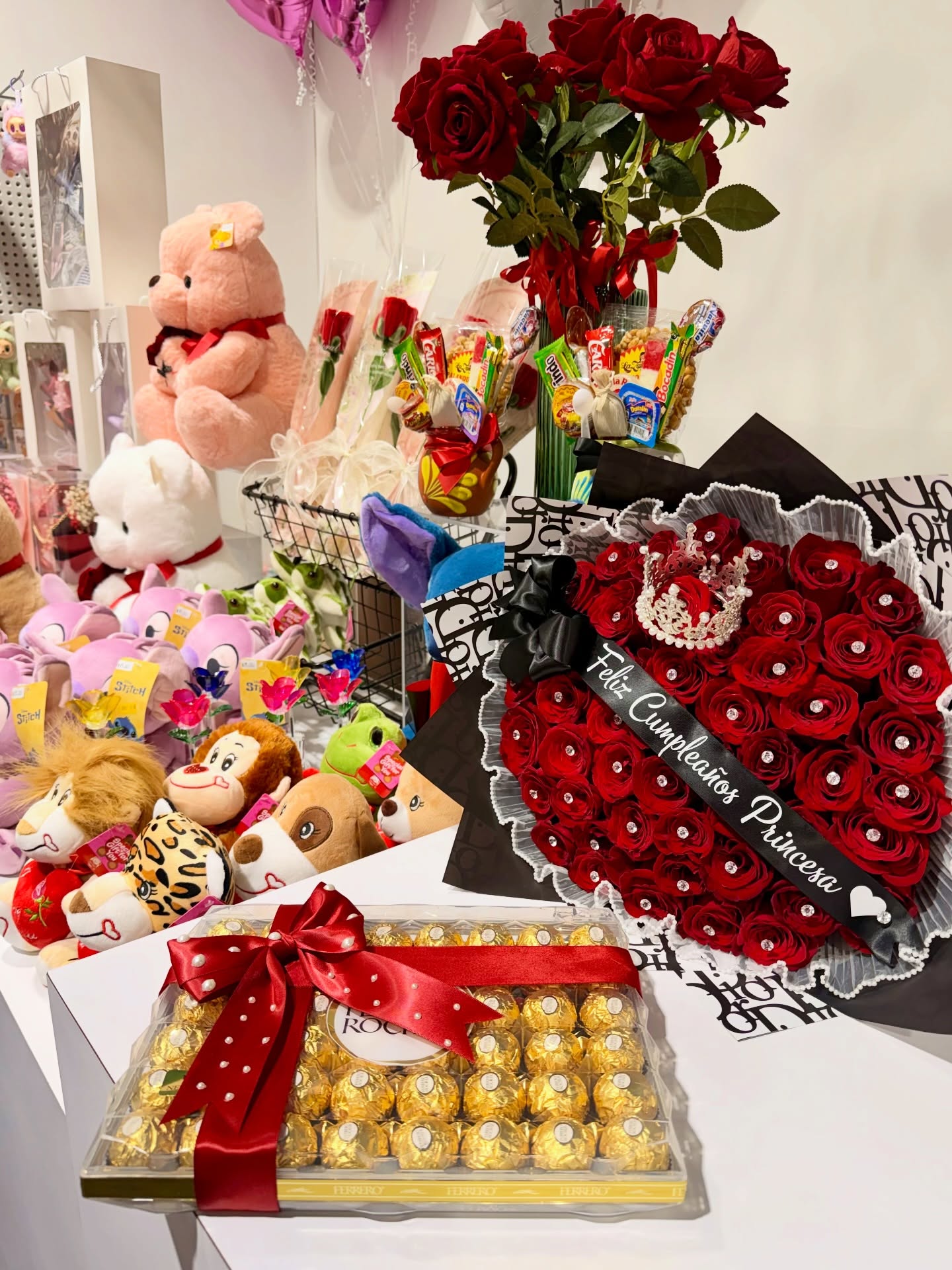 Gift shop display featuring a large box of Ferrero Rocher chocolates with a red bow and a lavish red rose bouquet with a tiara and "Feliz Cumpleaños Princesa" sash, surrounded by plush toys and candy arrangements.