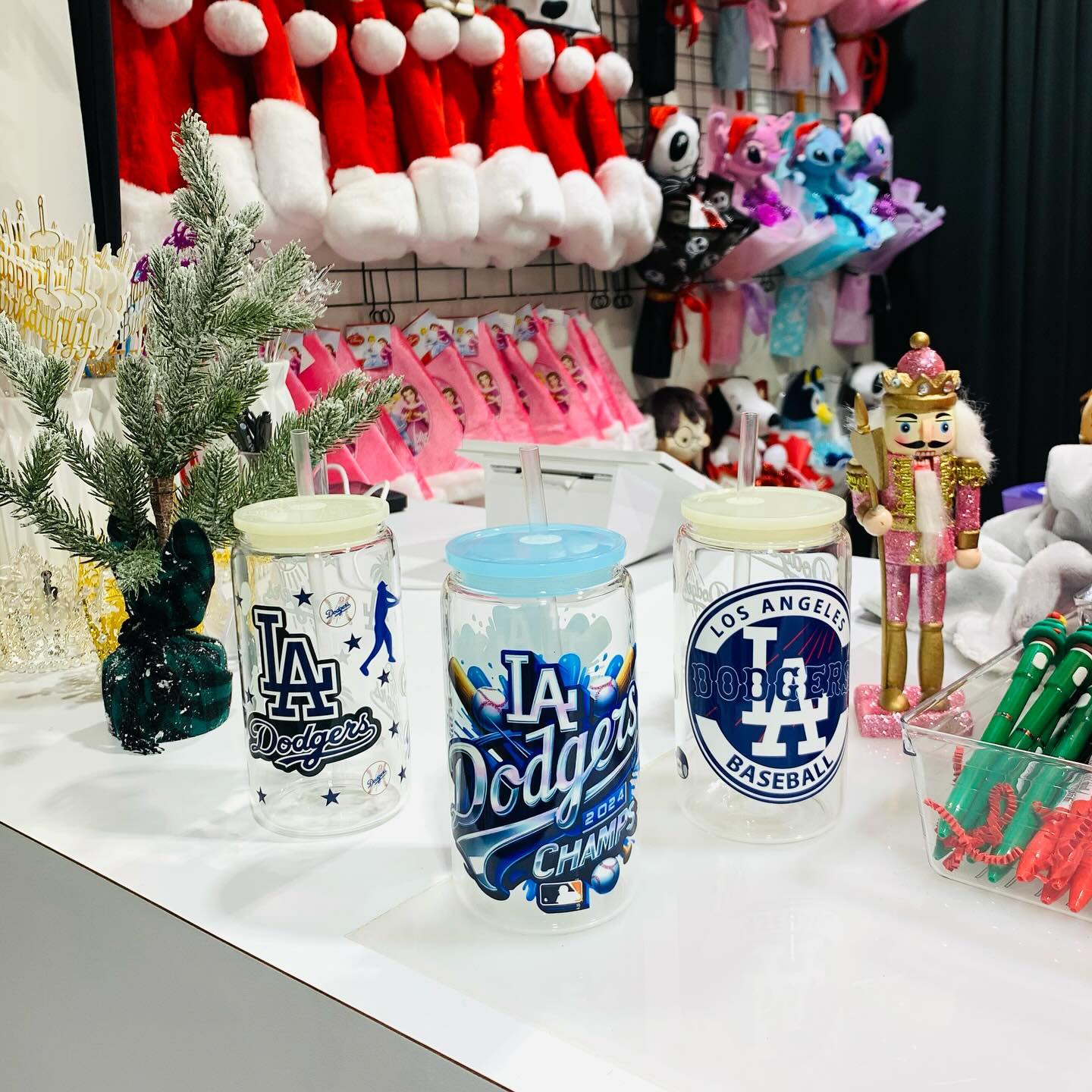 Three Los Angeles Dodgers themed clear glass tumblers with lids and straws, including a 2024 Champs design, displayed on a retail counter with Christmas holiday decorations in the background.
