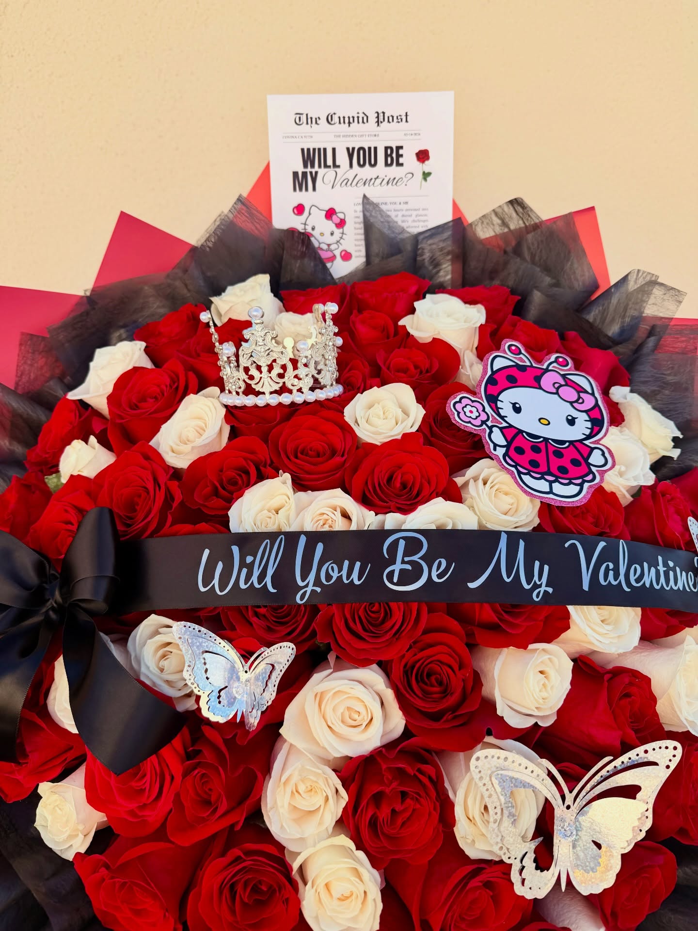 Valentine’s Day bouquet with red and white roses, Hello Kitty decoration, crown, butterflies, and “Will You Be My Valentine?” ribbon