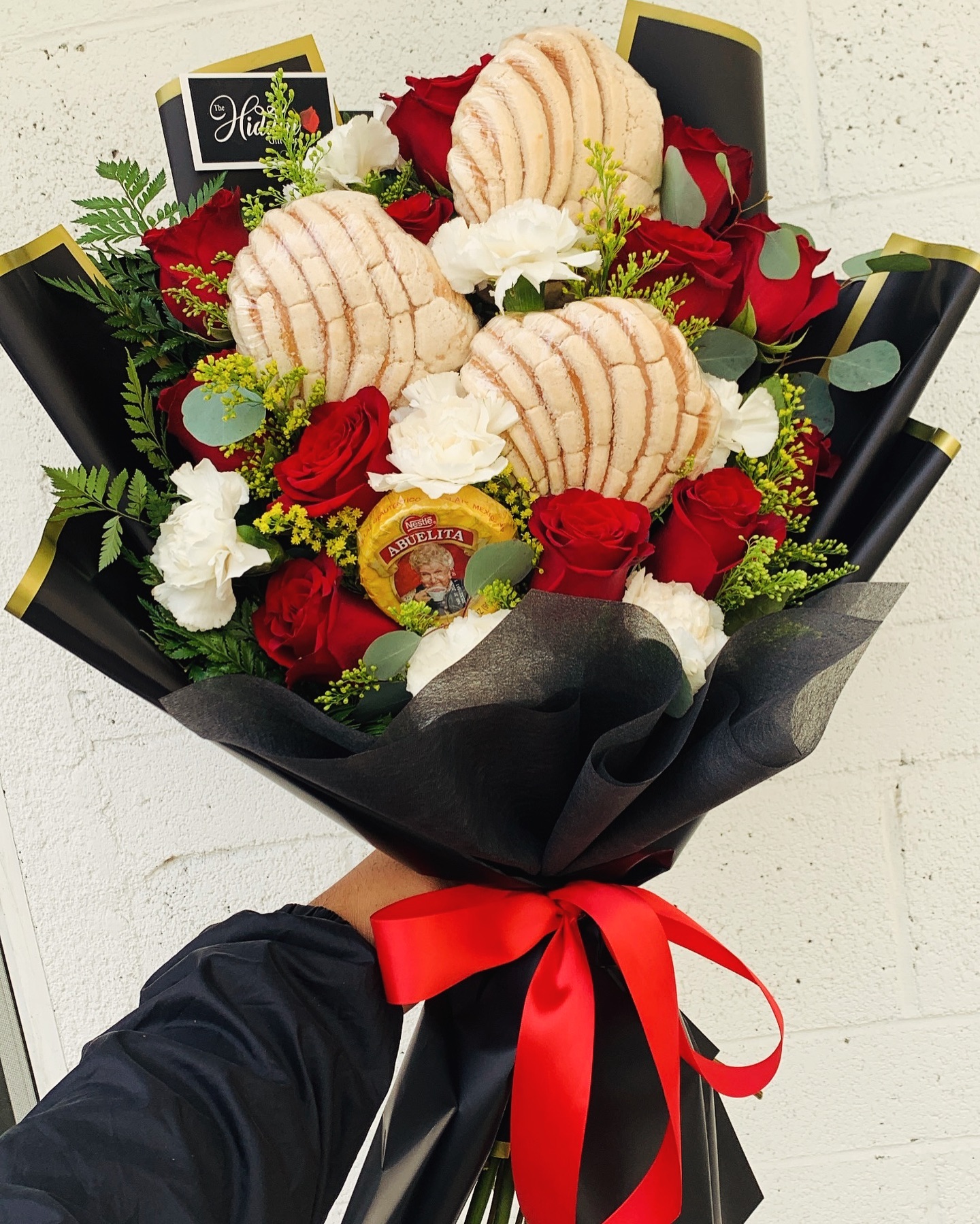 A creative Mexican-themed gift bouquet featuring red roses, white carnations, three traditional concha pastries (pan dulce), and a tablet of Abuelita hot chocolate, wrapped in black paper with a red ribbon.