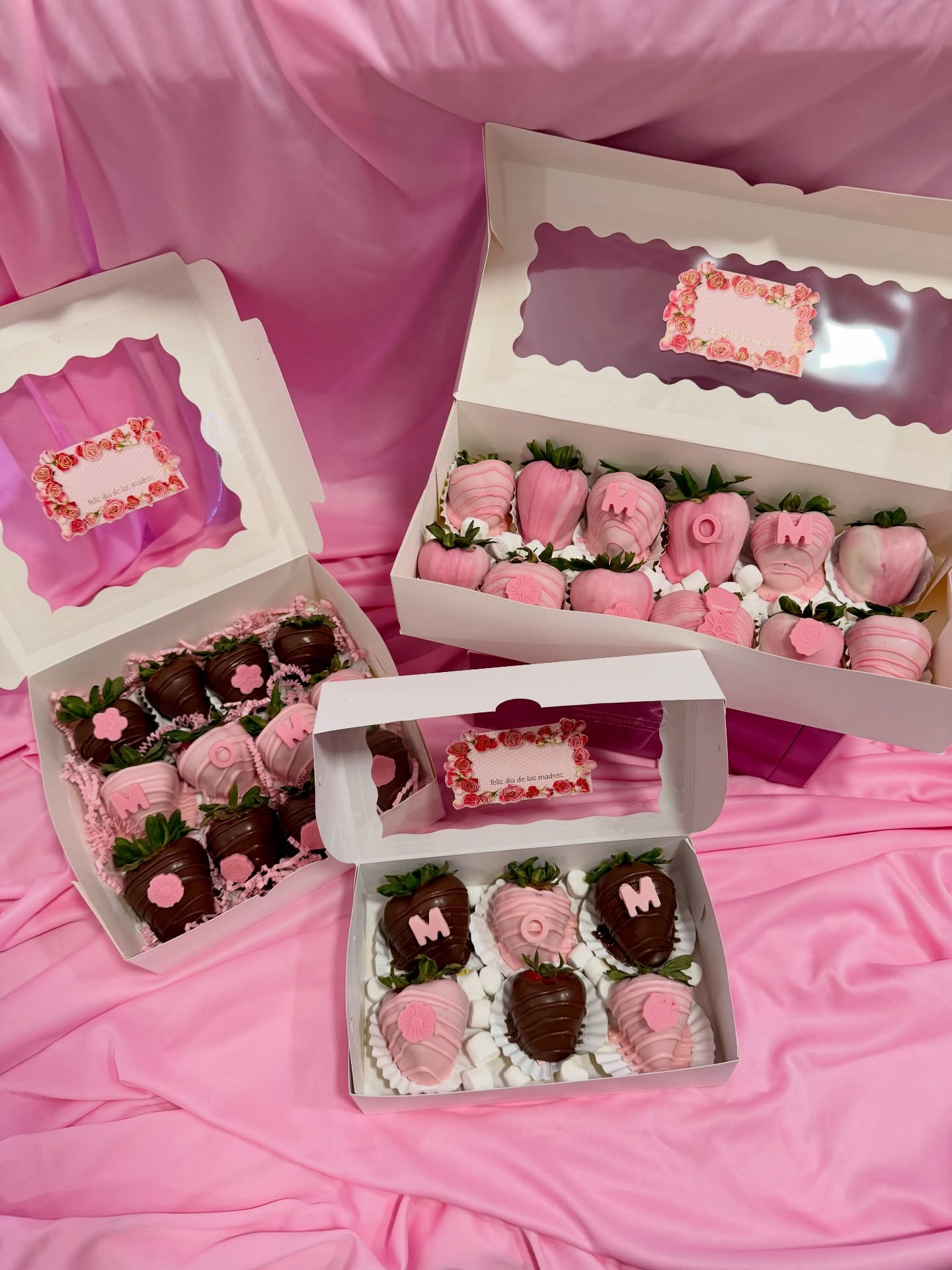 Three gift boxes of Mother's Day chocolate-covered strawberries decorated in pink and milk chocolate, featuring "MOM" lettering and floral accents, displayed on a draped pink fabric background.