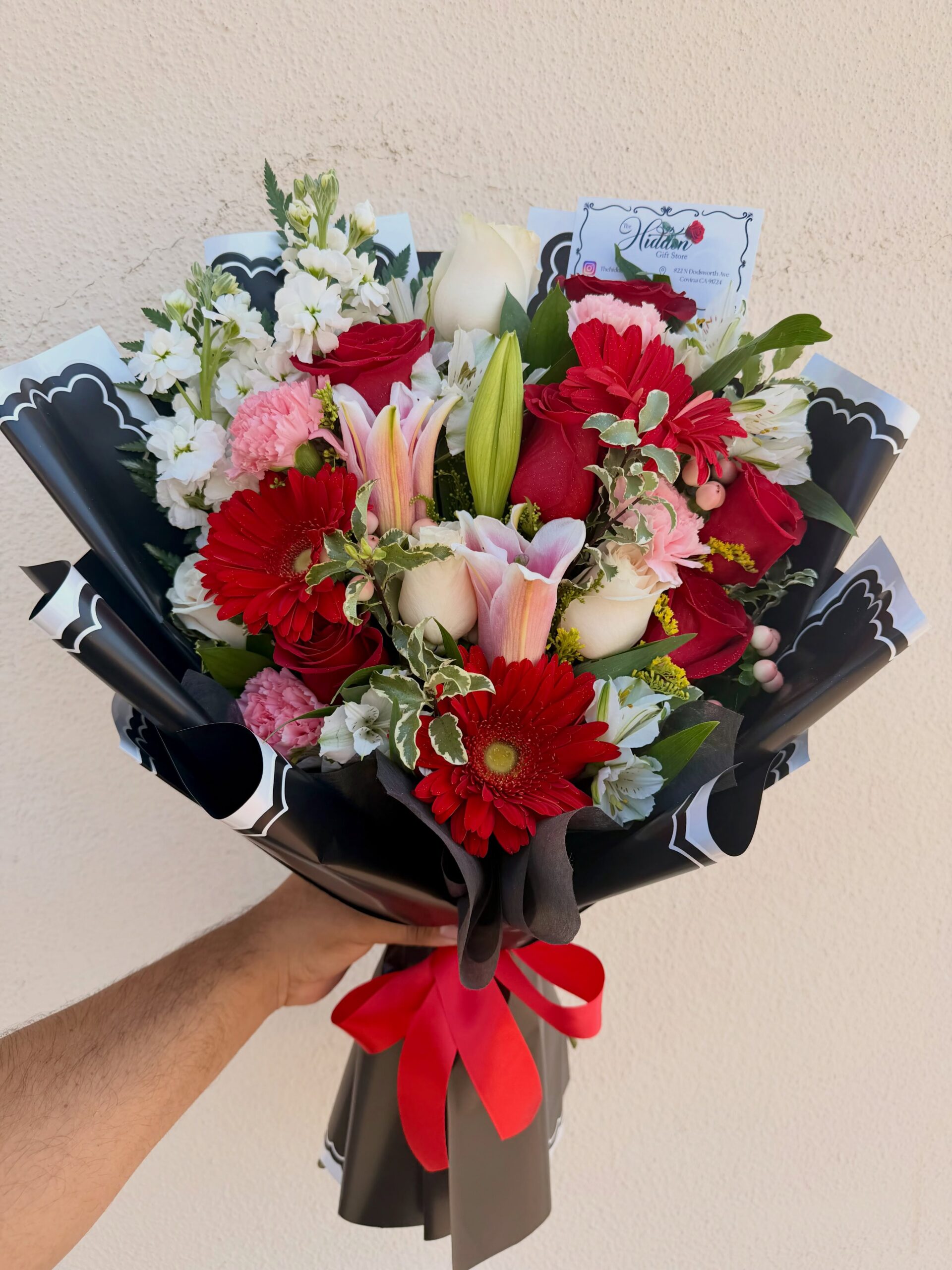 Red and white rose bouquet with pink lilies, red gerbera daisies, carnations, greenery, and black wrapping with a red ribbon